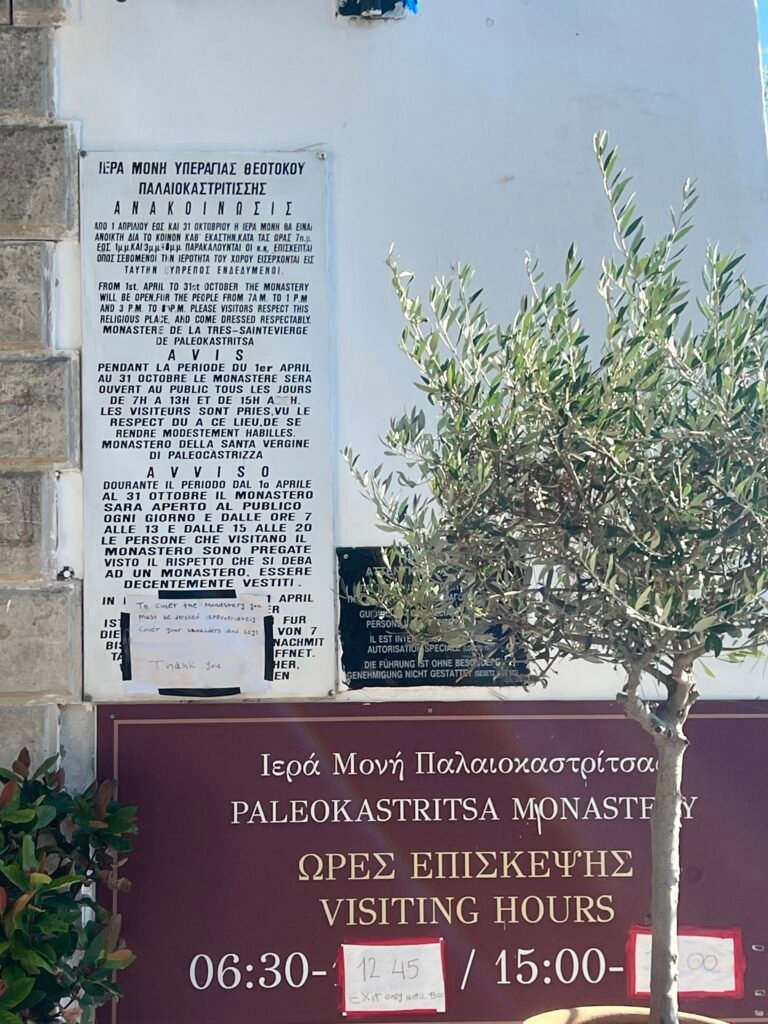 Paleokastritsa Monastery entrance notices showing visiting hours and a request to cover shoulders and legs